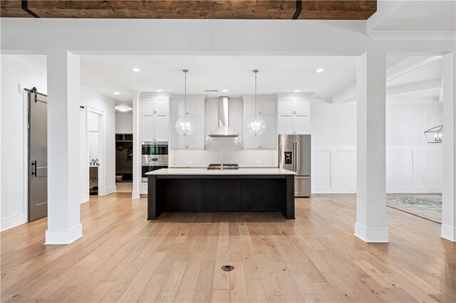 kitchen featuring light wood finished floors, wall chimney exhaust hood, appliances with stainless steel finishes, and a barn door