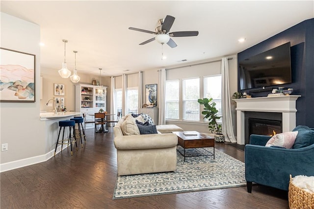 living area with dark wood-style floors, ceiling fan, a glass covered fireplace, and baseboards