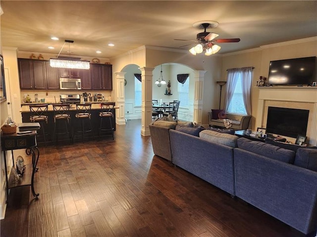 living room with arched walkways, ceiling fan, dark wood-style flooring, decorative columns, and crown molding