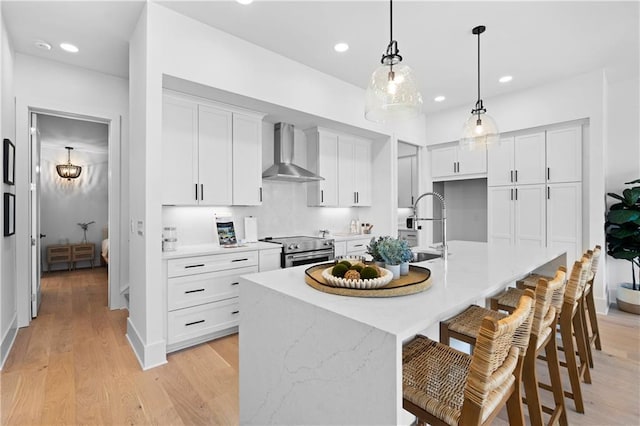 kitchen featuring a center island with sink, stainless steel electric range, wall chimney range hood, pendant lighting, and white cabinetry