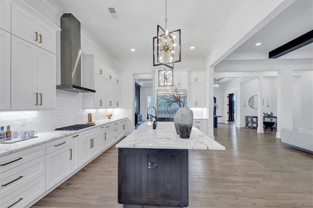 kitchen with wall chimney range hood, white cabinets, decorative light fixtures, and an island with sink