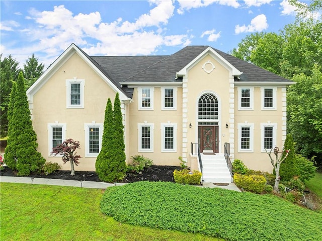 colonial-style house with roof with shingles, a front yard, and stucco siding