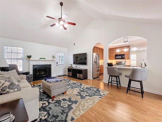 living room with high vaulted ceiling, arched walkways, light wood-style floors, a ceiling fan, and a high end fireplace