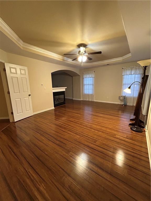 unfurnished living room with wood finished floors, ornamental molding, ceiling fan, a glass covered fireplace, and a raised ceiling