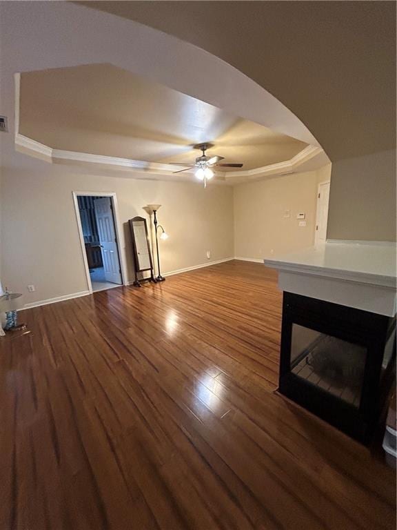 unfurnished living room with a tray ceiling, wood finished floors, arched walkways, baseboards, and ceiling fan