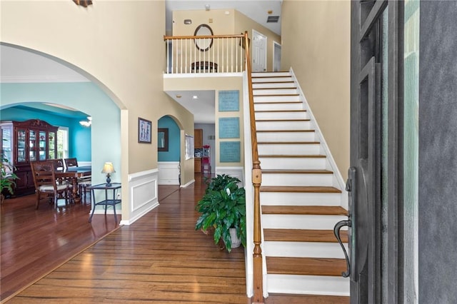 staircase featuring wood finished floors, visible vents, arched walkways, a towering ceiling, and wainscoting