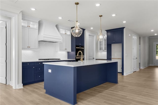 kitchen featuring blue cabinets, premium range hood, a kitchen island with sink, white cabinets, and light wood-type flooring