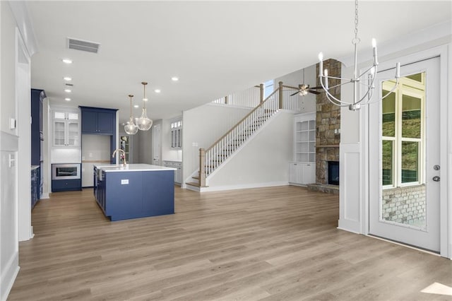 kitchen featuring a center island with sink, ceiling fan with notable chandelier, a stone fireplace, blue cabinets, and light hardwood / wood-style floors