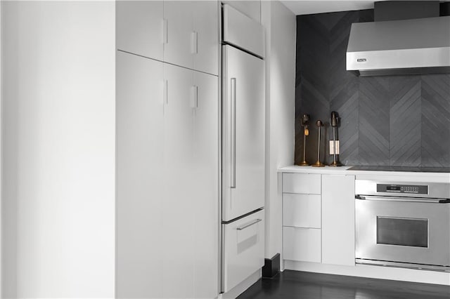 kitchen with wall chimney exhaust hood, stainless steel oven, paneled built in fridge, black electric stovetop, and white cabinets