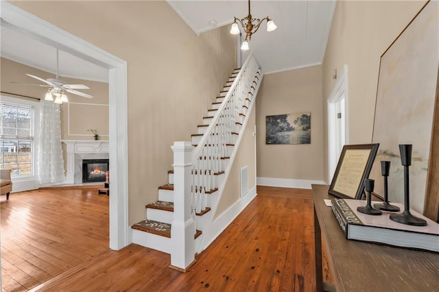 staircase with ceiling fan with notable chandelier, visible vents, hardwood / wood-style floors, a glass covered fireplace, and crown molding