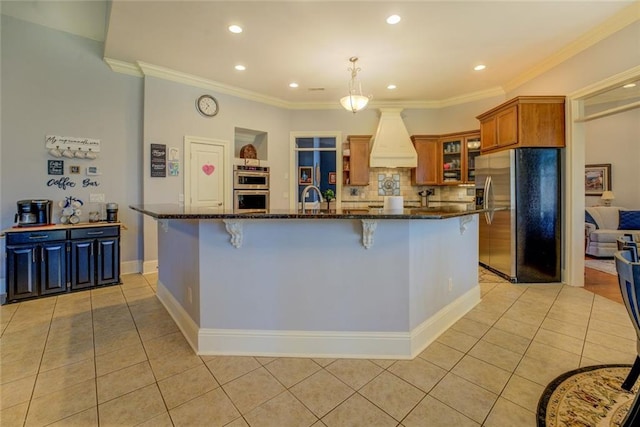 kitchen with appliances with stainless steel finishes, premium range hood, light tile patterned floors, brown cabinetry, and dark stone countertops