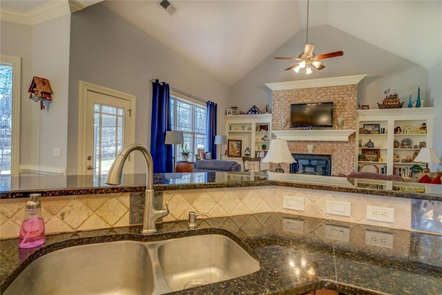 kitchen featuring a sink, ceiling fan, dark stone counters, a brick fireplace, and vaulted ceiling