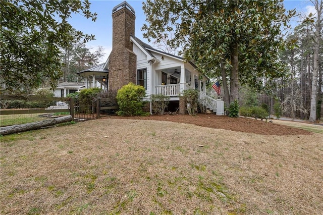 view of side of property featuring covered porch, a ceiling fan, a chimney, a lawn, and stairway