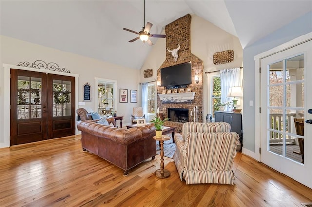 living room featuring a brick fireplace, light wood-style floors, high vaulted ceiling, french doors, and a ceiling fan
