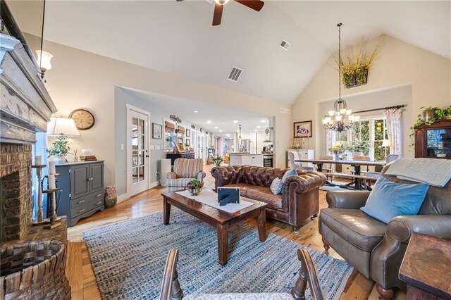 living area featuring visible vents, a fireplace, high vaulted ceiling, light wood-style flooring, and ceiling fan with notable chandelier