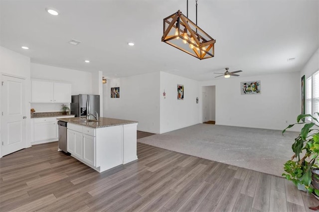 kitchen featuring light wood-type flooring, appliances with stainless steel finishes, an island with sink, dark stone counters, and white cabinets