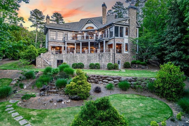 back of property at dusk featuring a balcony, stone siding, a chimney, stairs, and a yard