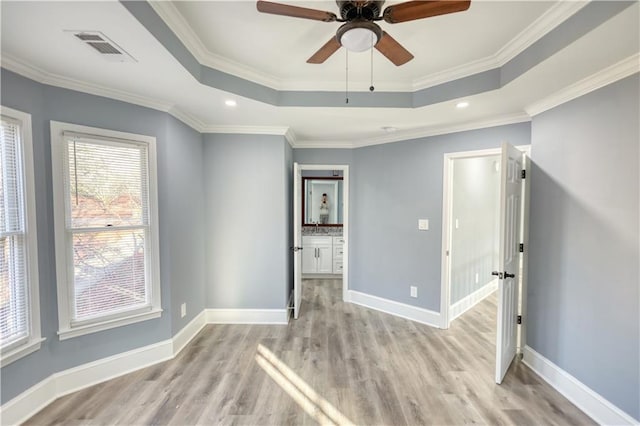 empty room with visible vents, light wood-style flooring, a tray ceiling, crown molding, and baseboards