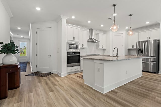 kitchen with appliances with stainless steel finishes, wall chimney exhaust hood, a sink, light wood finished floors, and tasteful backsplash