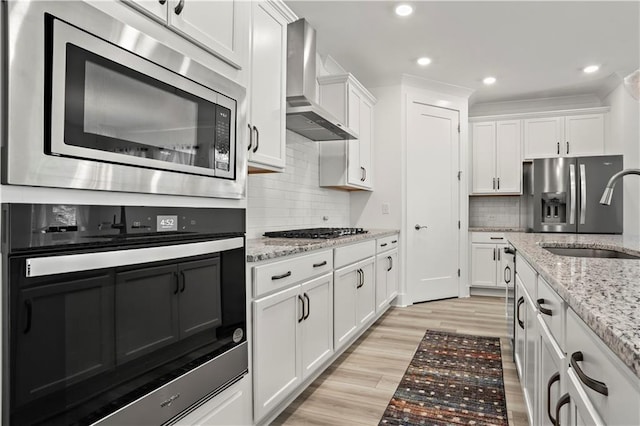 kitchen with appliances with stainless steel finishes, wall chimney exhaust hood, a sink, white cabinetry, and recessed lighting