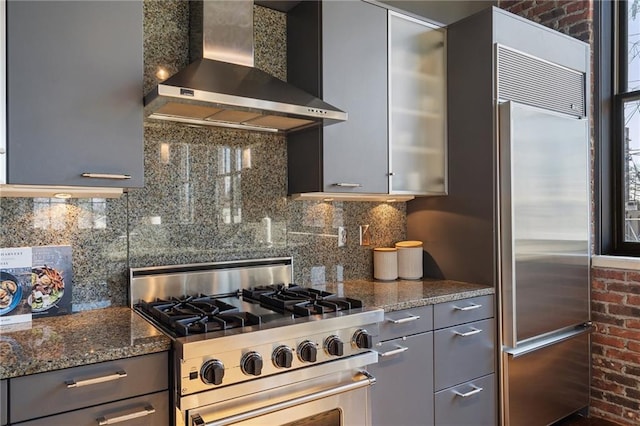 kitchen with gray cabinets, dark stone countertops, high quality appliances, decorative backsplash, and wall chimney exhaust hood