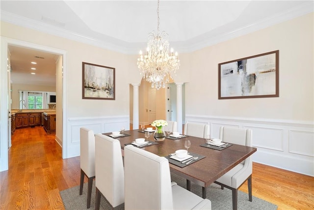 dining space with a chandelier, a wainscoted wall, light wood-style floors, decorative columns, and crown molding