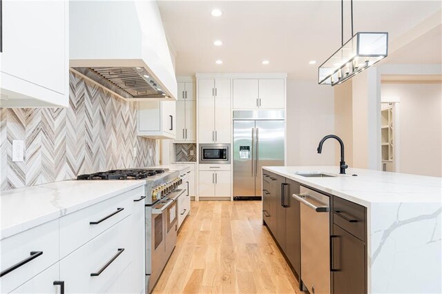 kitchen with built in appliances, a center island with sink, custom exhaust hood, light wood-type flooring, and white cabinets