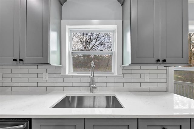 kitchen with light stone counters, gray cabinets, a sink, dishwasher, and backsplash