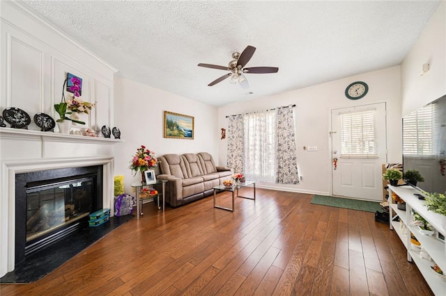 living room featuring a glass covered fireplace, dark wood-type flooring, a textured ceiling, baseboards, and a ceiling fan