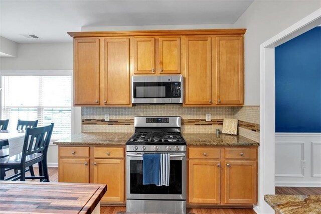 kitchen with stainless steel appliances, stone counters, light wood finished floors, backsplash, and wainscoting