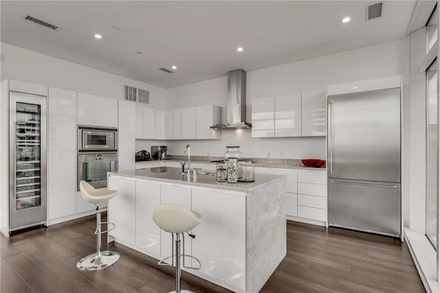 kitchen featuring white cabinetry, wall chimney exhaust hood, built in appliances, and a center island with sink