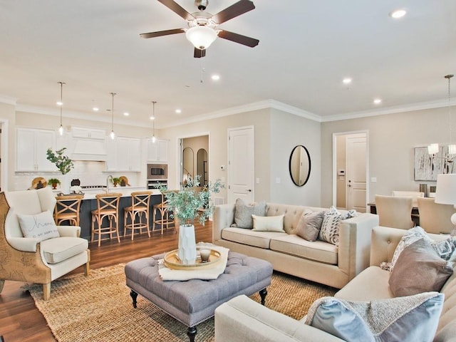 living room featuring dark wood finished floors, ornamental molding, ceiling fan with notable chandelier, and recessed lighting