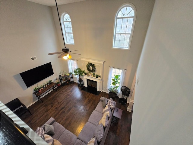 living room featuring visible vents, a towering ceiling, a fireplace with flush hearth, dark wood-type flooring, and ceiling fan