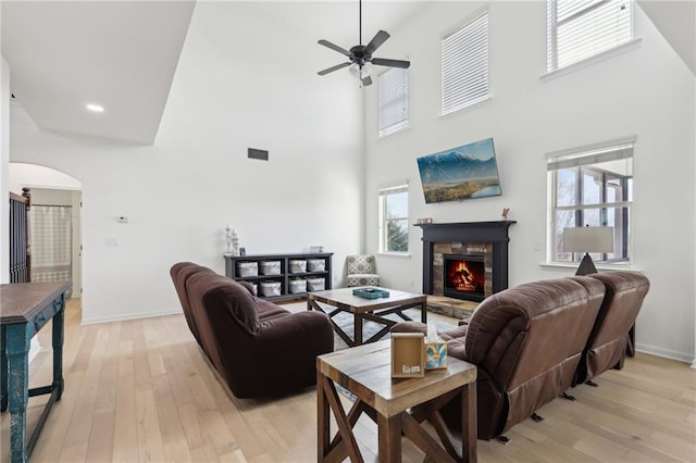 living room featuring a stone fireplace, light hardwood / wood-style floors, ceiling fan, and a towering ceiling