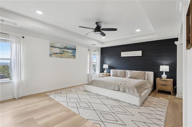 bedroom featuring ceiling fan, wooden walls, light wood-type flooring, and a tray ceiling