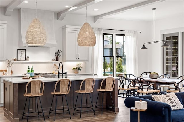 kitchen featuring pendant lighting, a breakfast bar area, a kitchen island with sink, beam ceiling, and white cabinets