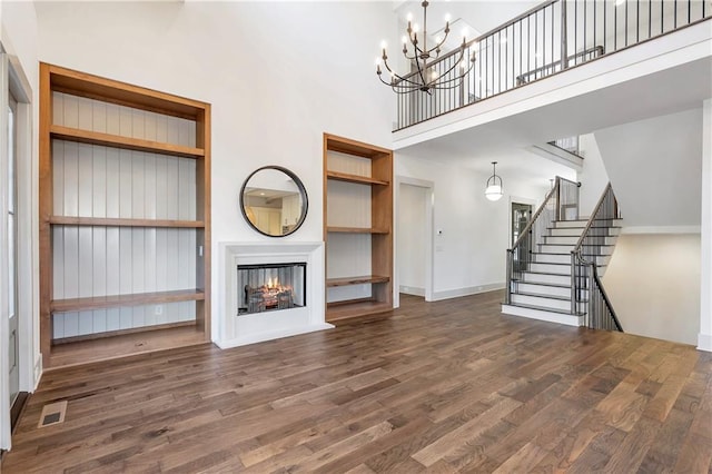 unfurnished living room featuring dark wood-type flooring, built in features, an inviting chandelier, a towering ceiling, and a multi sided fireplace