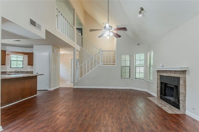 unfurnished living room with high vaulted ceiling, a ceiling fan, dark wood-type flooring, a tiled fireplace, and stairway