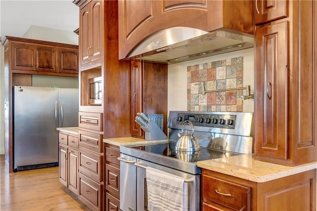 kitchen featuring stainless steel appliances, ventilation hood, light stone countertops, light wood finished floors, and brown cabinetry