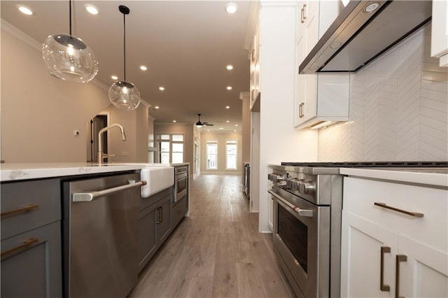 kitchen featuring wall chimney exhaust hood, white cabinetry, decorative light fixtures, ornamental molding, and appliances with stainless steel finishes