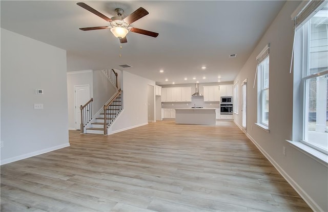 unfurnished living room with light wood-style flooring, visible vents, baseboards, stairs, and a ceiling fan