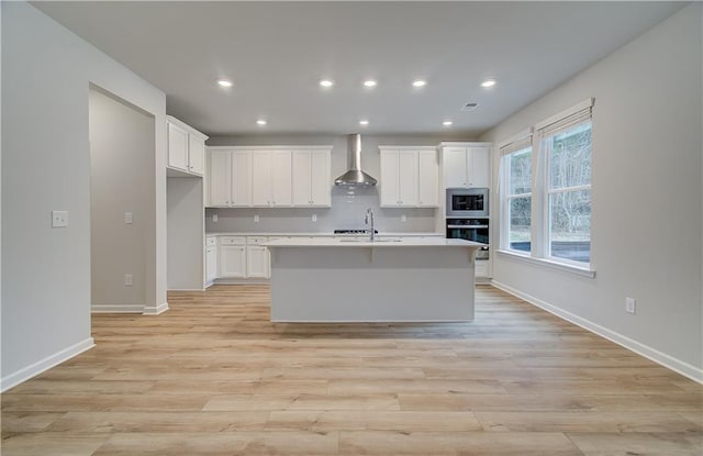 kitchen with light countertops, built in microwave, wall chimney range hood, a kitchen island with sink, and oven