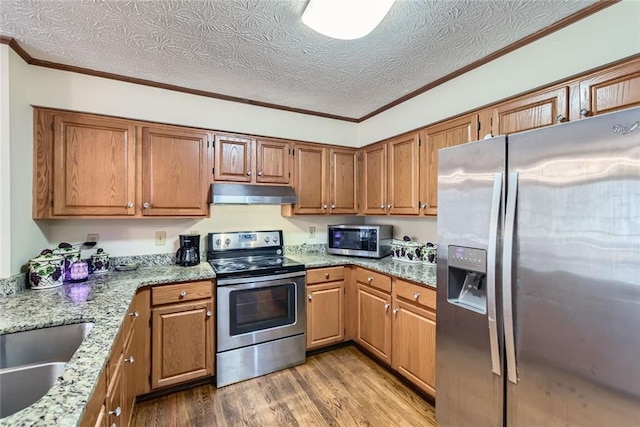 kitchen featuring light stone countertops, wood finished floors, ornamental molding, under cabinet range hood, and appliances with stainless steel finishes