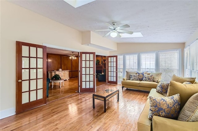 living area featuring vaulted ceiling with skylight, light wood-style flooring, ceiling fan, french doors, and a textured ceiling