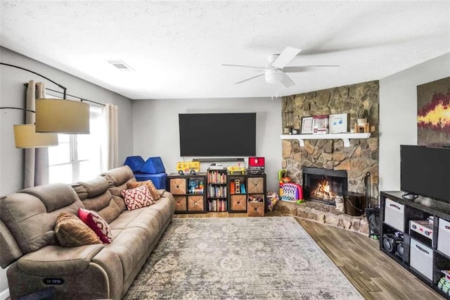 living area with visible vents, a ceiling fan, a stone fireplace, a textured ceiling, and wood finished floors