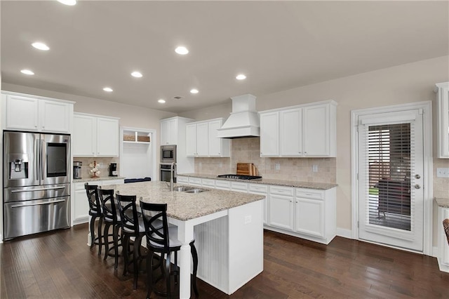 kitchen featuring premium range hood, appliances with stainless steel finishes, white cabinetry, an island with sink, and light stone countertops