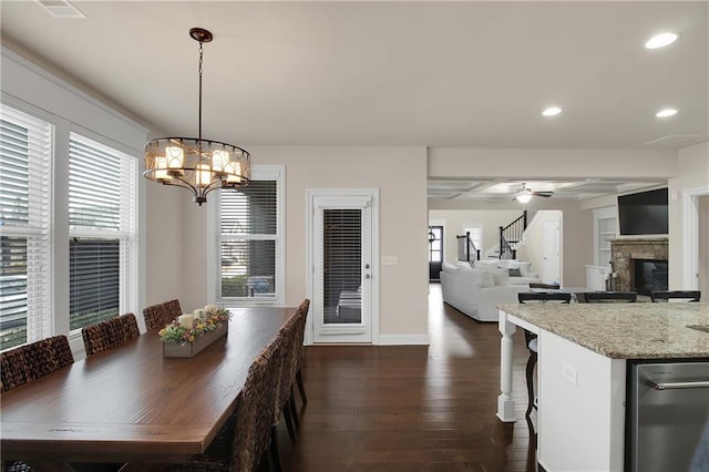 dining area with a stone fireplace, dark wood-type flooring, and ceiling fan with notable chandelier