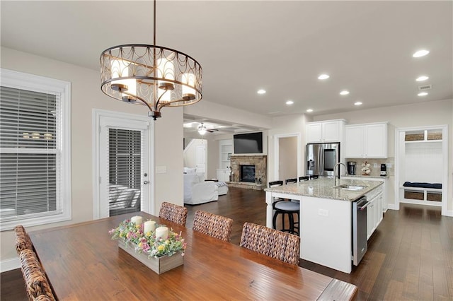 dining area featuring a stone fireplace, sink, ceiling fan with notable chandelier, and dark hardwood / wood-style floors