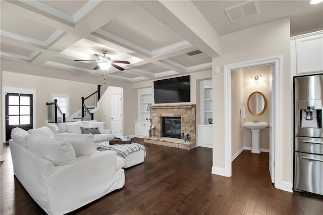 living room featuring a stone fireplace, dark hardwood / wood-style floors, coffered ceiling, ceiling fan, and beam ceiling