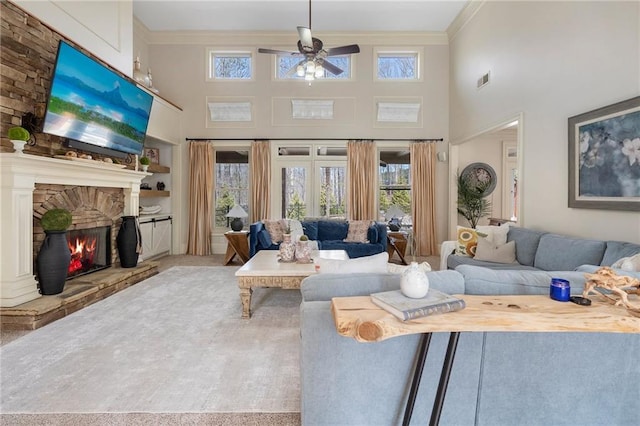 carpeted living room with a high ceiling, a stone fireplace, a ceiling fan, and crown molding